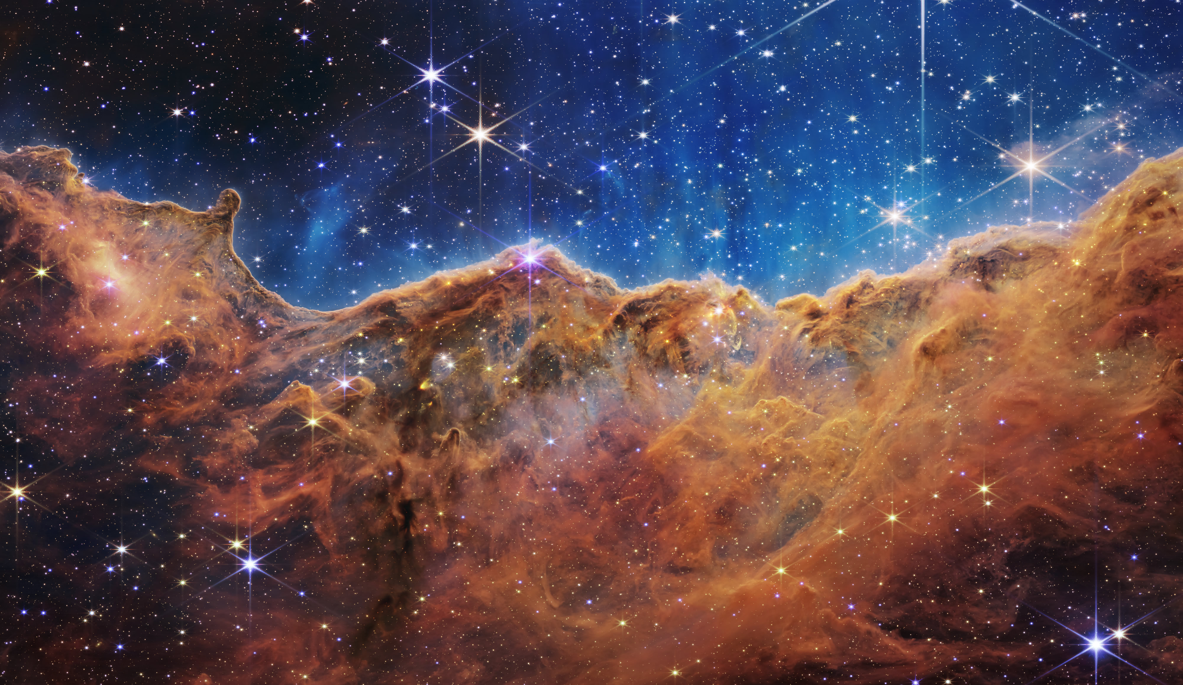 Image from the James Webb Space Telescope of a star forming region in Carina Nebula. The bottom 75% of the image is orange clouds that look like cliffs and mountains. In the top 25% is a blue and black canvas with stars and other white specs.