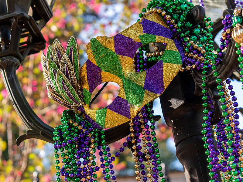 A Mardi Gras mask and beads hang off of a street light. The mask and the beads are gold, purple, and green.