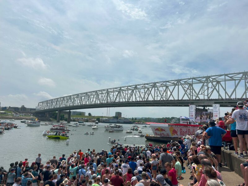 A large crowd stands on the shore and in boats along the Ohio River while they watch teams compete in Red Bull Flugtag in Cincinnati, Ohio. Engineering Human-Powered Flight: What Is Red Bull Flugtag?